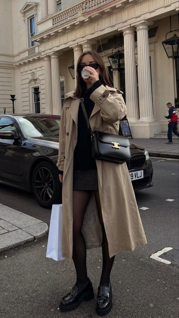 Stylish woman in trench coat sipping coffee, standing by car in urban street. Fashionable city lifestyle scene.