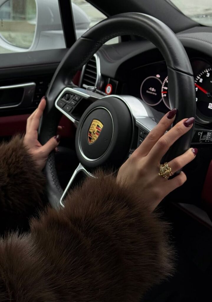 Hands on a luxury car steering wheel, wearing fur coat and gold ring, showcasing elegance and style.