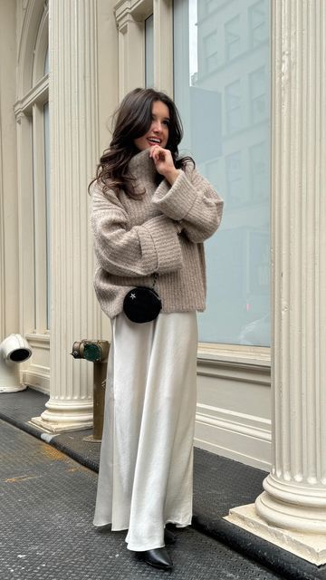 Woman in a cozy sweater and long skirt poses stylishly outside a building, holding a black purse.