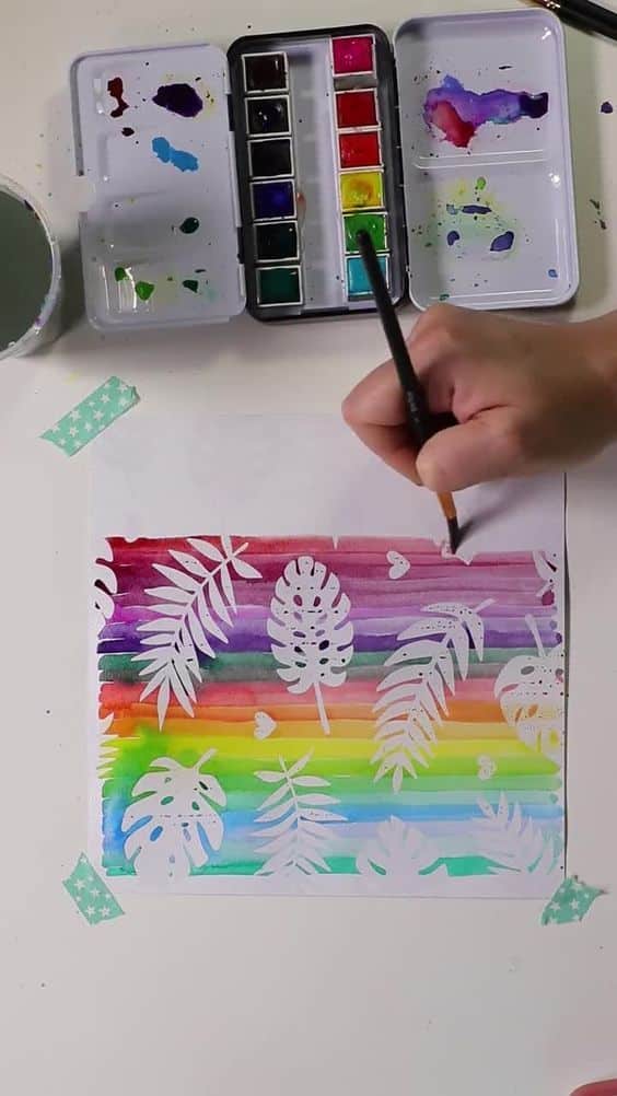 Hand painting a rainbow watercolor background with leaf patterns using a paintbrush and palette.