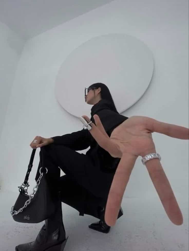 Person in stylish black outfit with rings and handbag, posing creatively against a minimal white background.