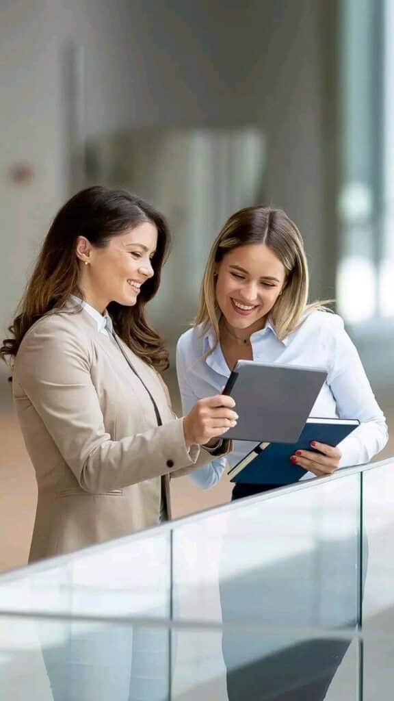 Two professional women happily discussing work on a tablet in a modern office setting.