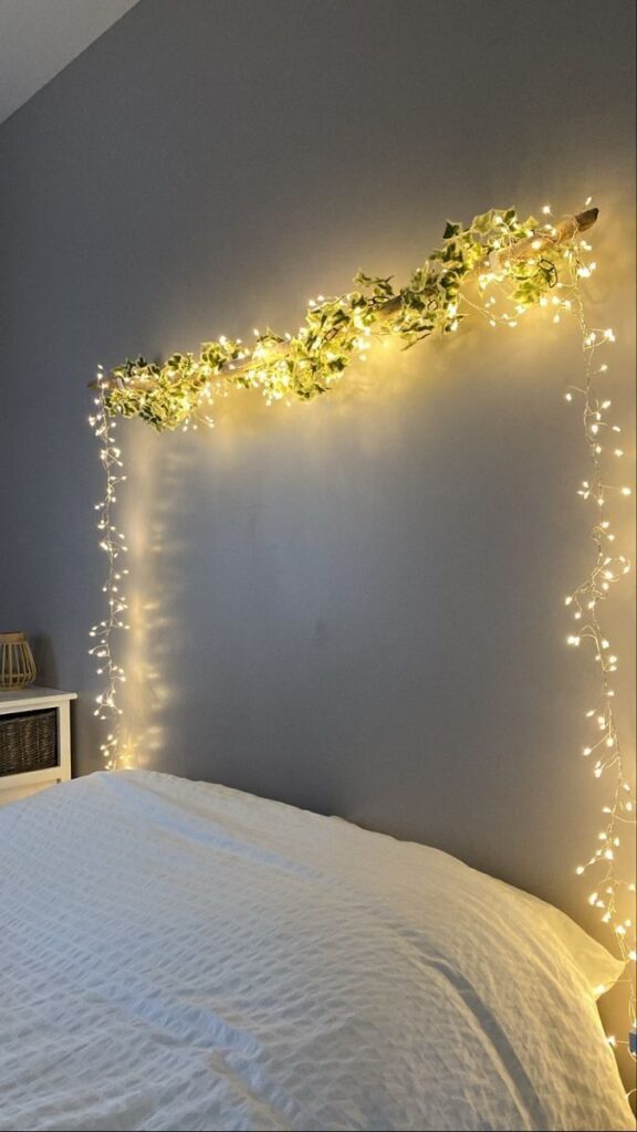 Cozy bedroom decor with string lights and greenery on a gray wall above a white bed.
