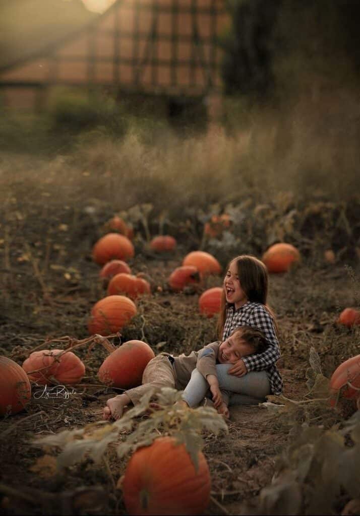 Two children laughing and playing among pumpkins in a field during autumn sunset.