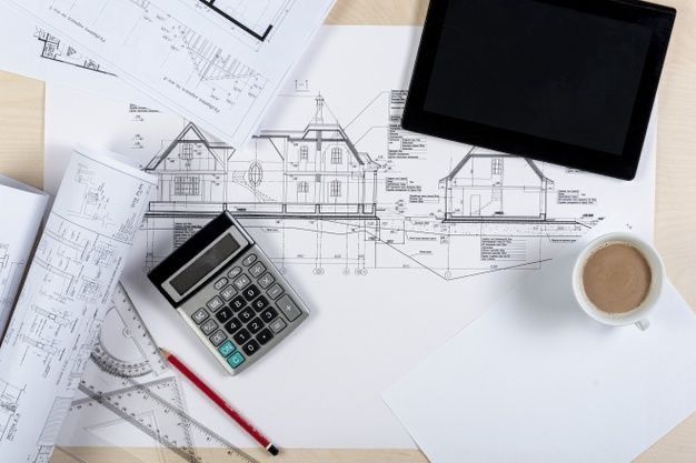 Architectural blueprints with a calculator, coffee, tablet, and drafting tools on a desk.