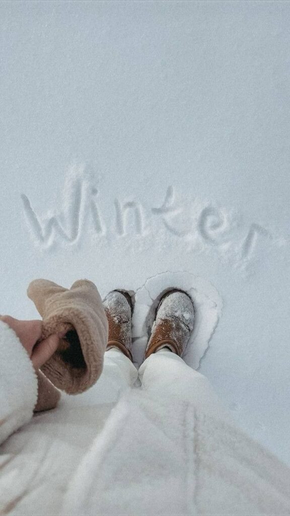 Person wearing winter boots in snow with 'winter' written on ground, holding a cozy hat.