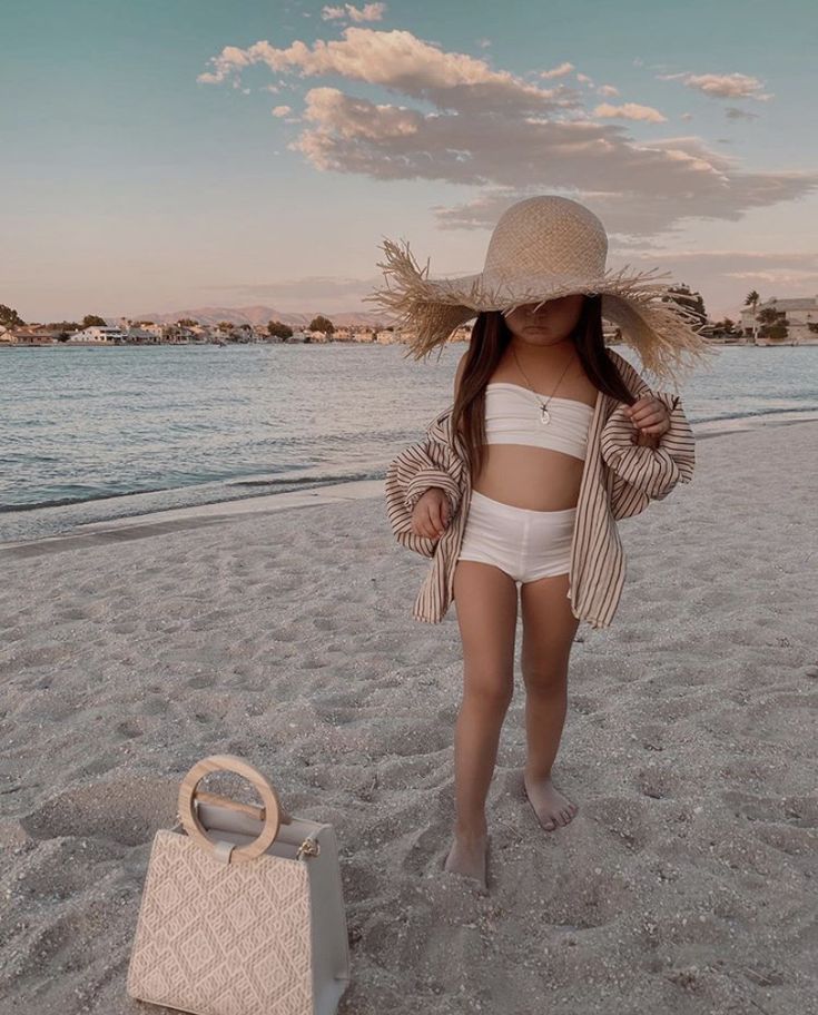 Child in beachwear and straw hat walking on a sandy beach near the water, with a stylish handbag nearby.
