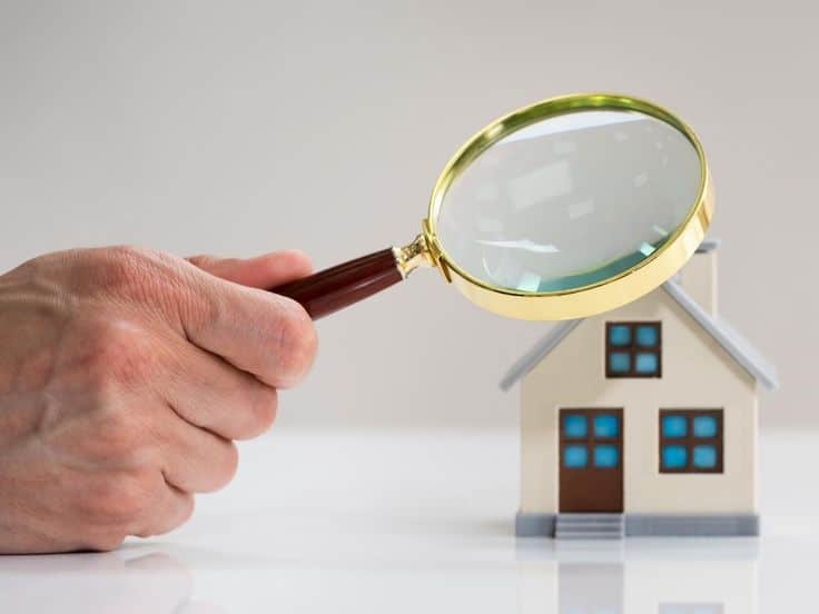 Hand holding magnifying glass examining small house model, symbolizing detailed home inspection or property search.