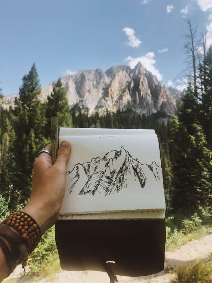 Hand holding a sketchbook with a mountain drawing, real mountains in the background under a blue sky.