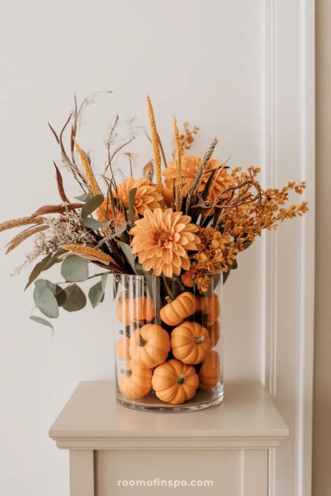 Elegant fall floral arrangement with mini pumpkins and orange blooms in a glass vase on a table.