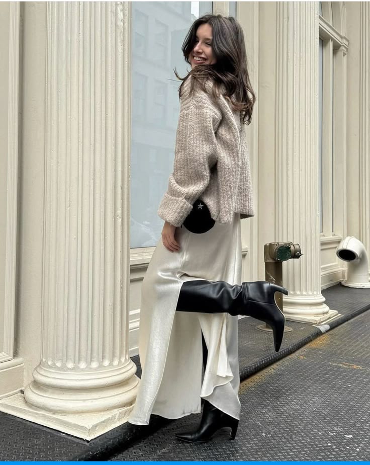 Woman in stylish winter outfit, cream skirt, and black boots, posing joyfully on city street with classical architecture.