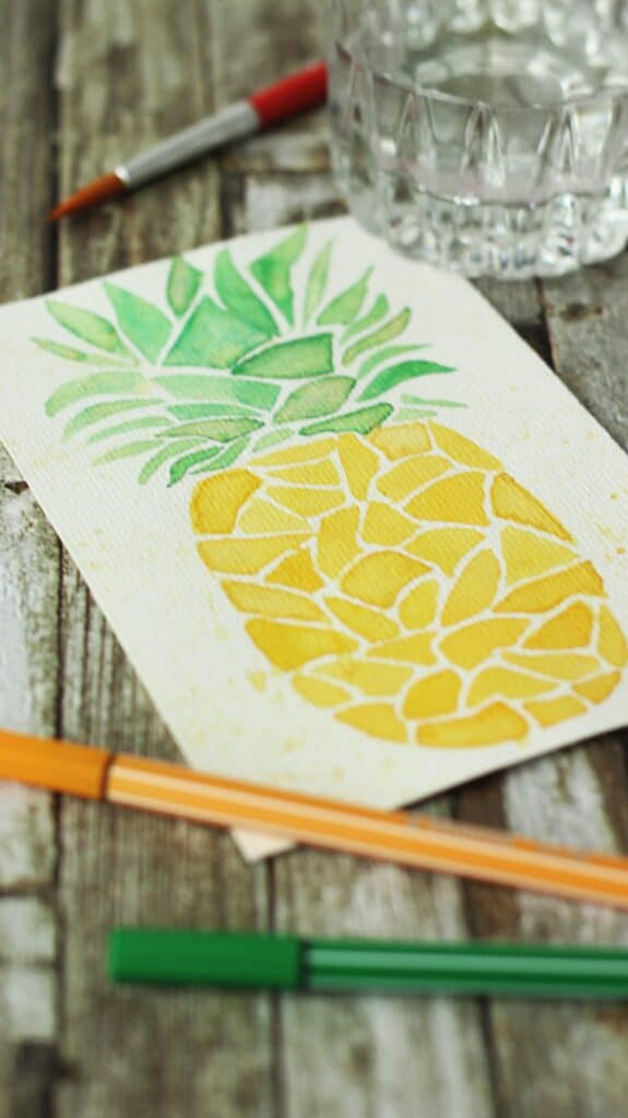 Watercolor pineapple art on rustic wood table with colored markers and a glass of water.