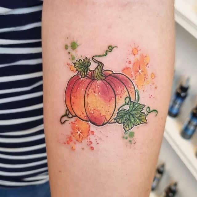 Colorful pumpkin tattoo on arm with vine details, surrounded by splashes of orange and green ink.