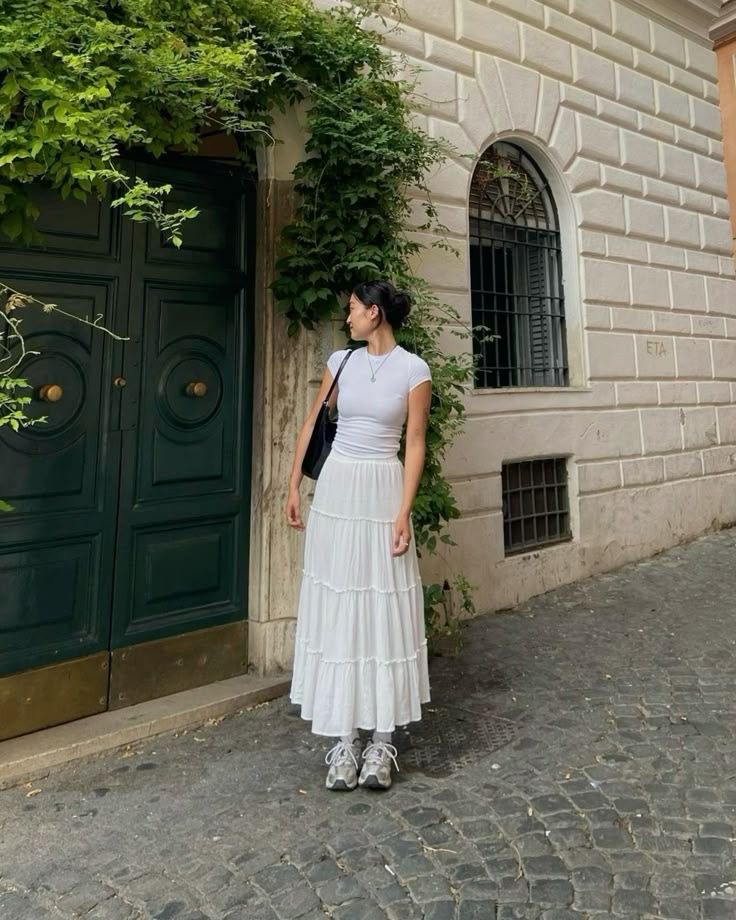 Woman in a white dress stands on a cobblestone street by a vine-covered doorway and arched window.