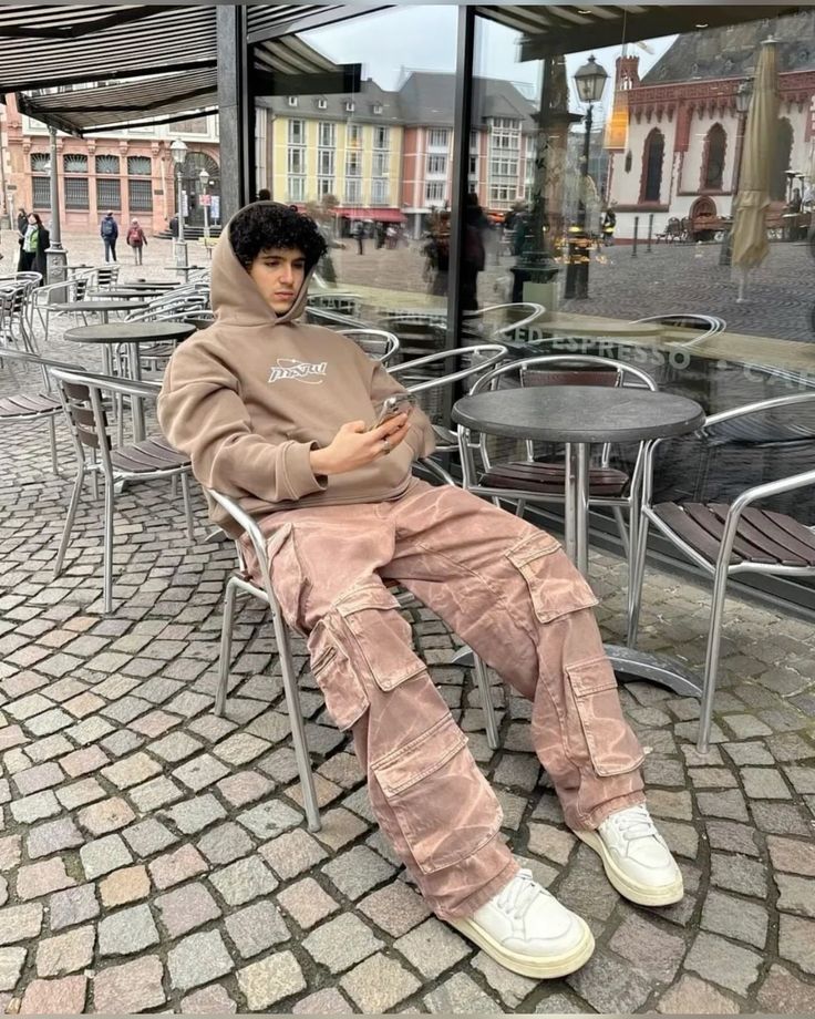 Young man in casual streetwear sits outside a cafe, using his phone on a cobblestone street with historic buildings.