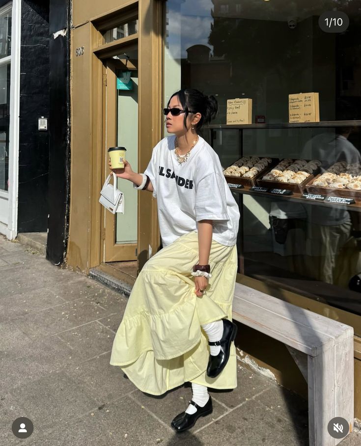 Fashionable woman in street style attire, seated outside a bakery, holding coffee and wearing sunglasses and a long skirt.