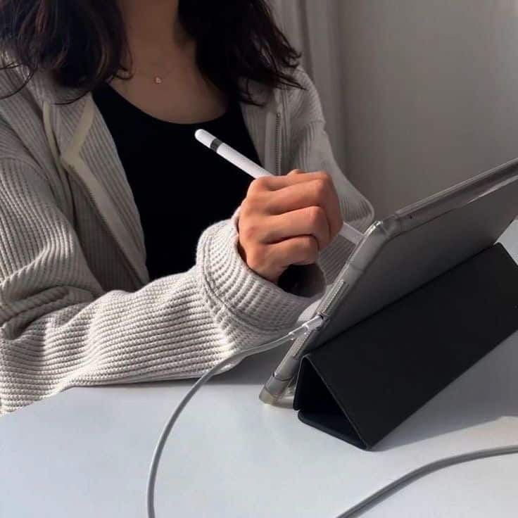 Person using a stylus on a tablet at a desk, wearing a cozy sweater in natural light.