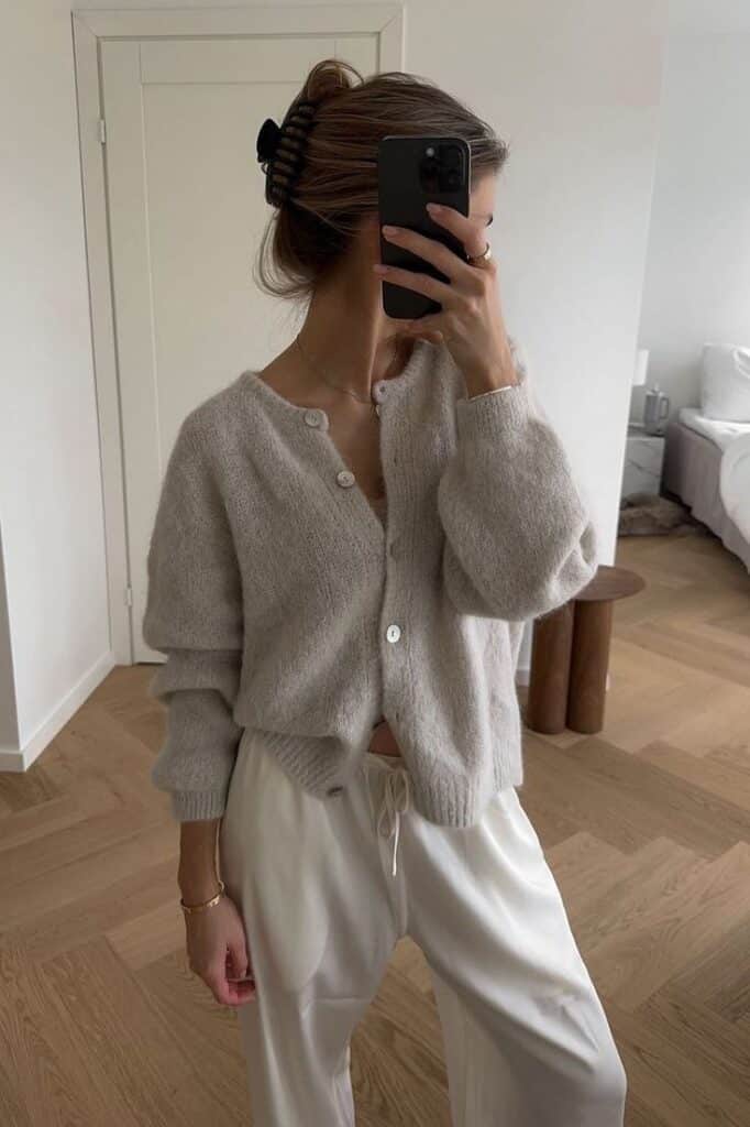 Woman in cozy beige cardigan and white pants taking selfie in stylish room.