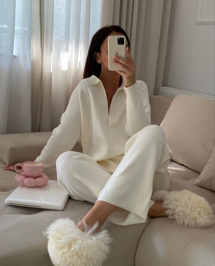 Cozy woman in white loungewear and fluffy slippers, taking a mirror selfie with a smartphone on a comfy sofa.