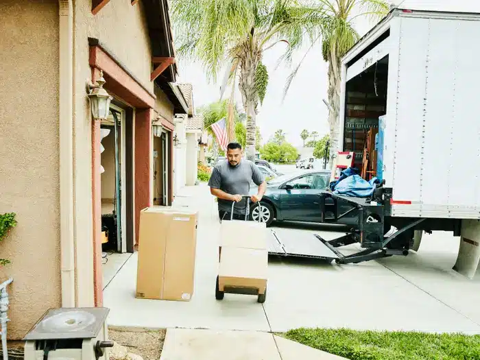 5f3e95b3cdf25a001e84e6e7 Man moving boxes into a house from a truck with a dolly on a sunny day. | Sky Rye Design Man moving boxes into a house from a truck with a dolly on a sunny day.
