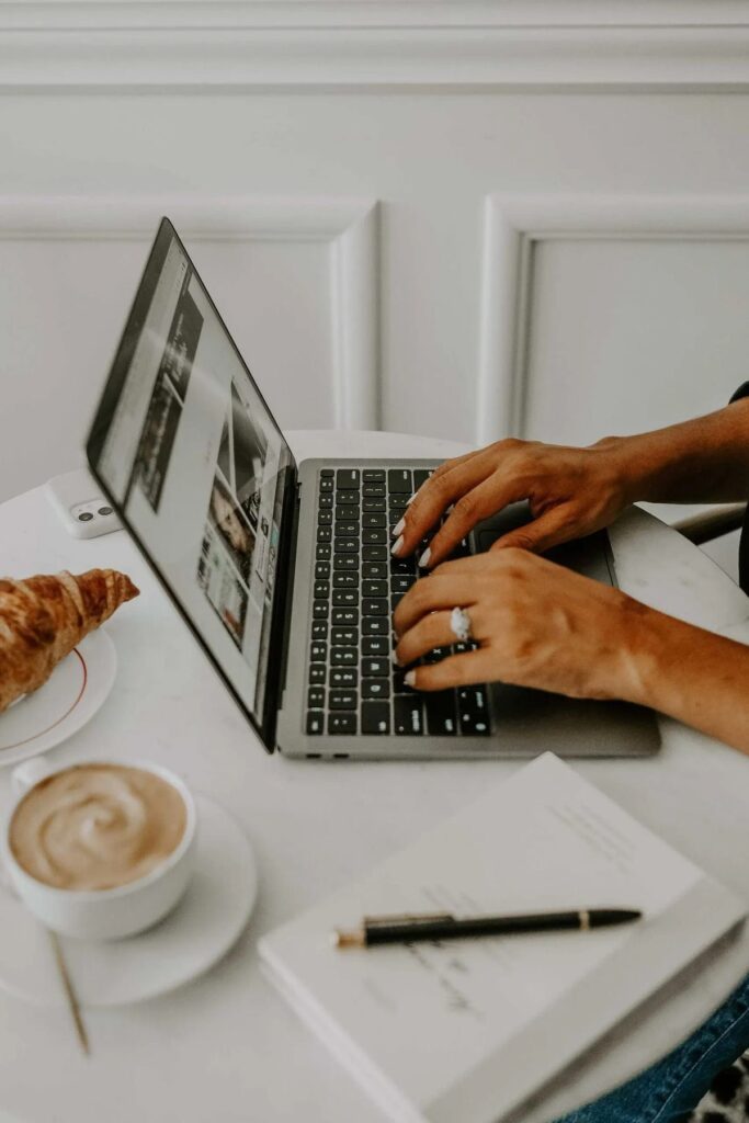 Person typing on laptop at cafe table with coffee, croissant, and notebook nearby.