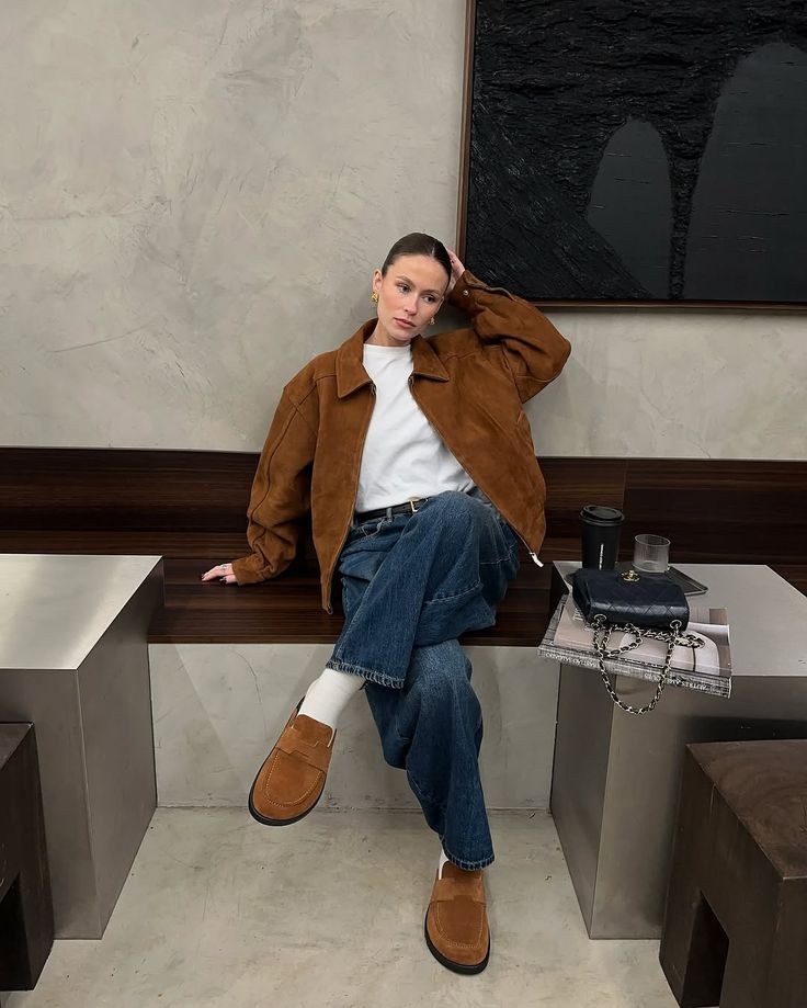 Person in brown jacket and jeans sitting on a bench with modern decor, holding a pose for a stylish, casual look.