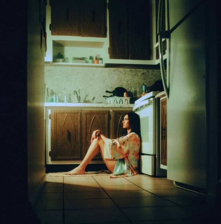 Woman sitting on kitchen floor in soft light, reflecting, surrounded by wooden cabinets and appliances.