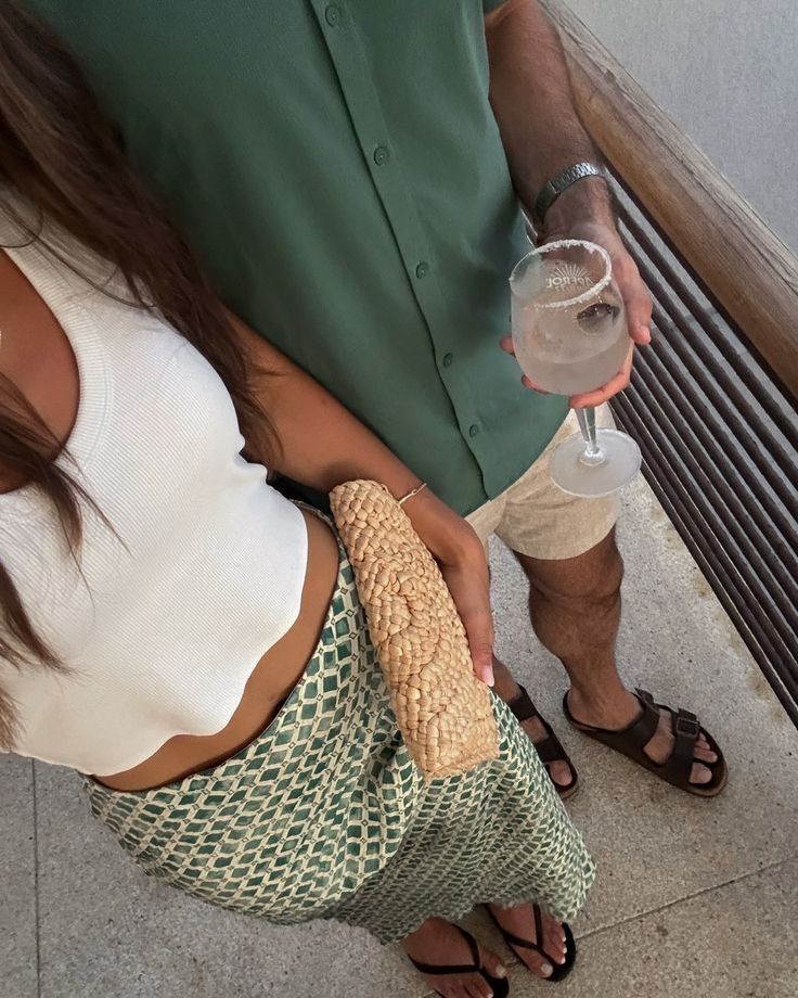 Couple in summer attire, woman with woven purse, man holding a cocktail, standing on a balcony with natural light.