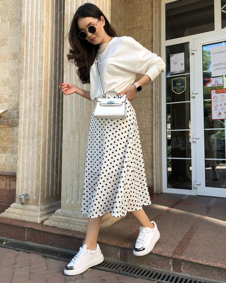 Fashionable woman in polka dot skirt, white top, sneakers, and sunglasses holding a mini bag outside a building.