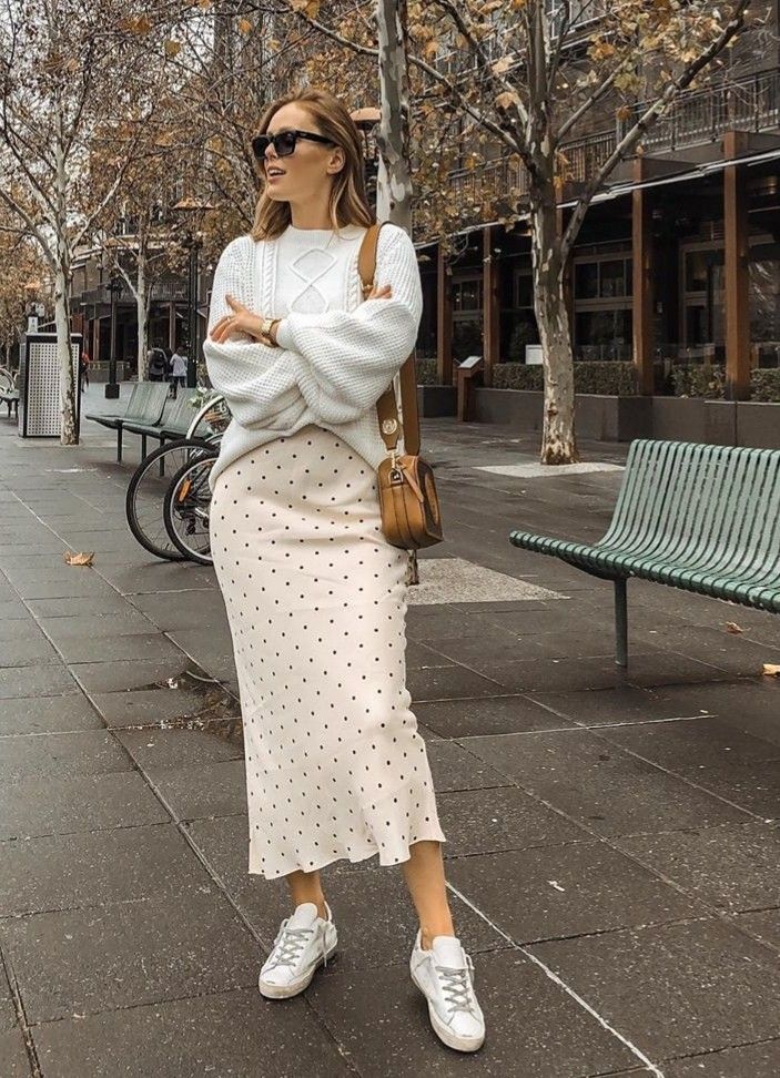 Woman in chic winter outfit with polka dot skirt and sweater, standing in urban setting with a brown bag and sunglasses.