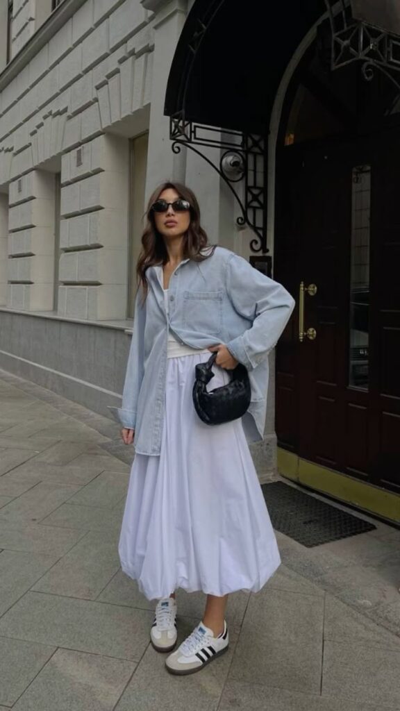 Fashionable woman in a denim shirt, white skirt, and sneakers, holding a black handbag on a city sidewalk.