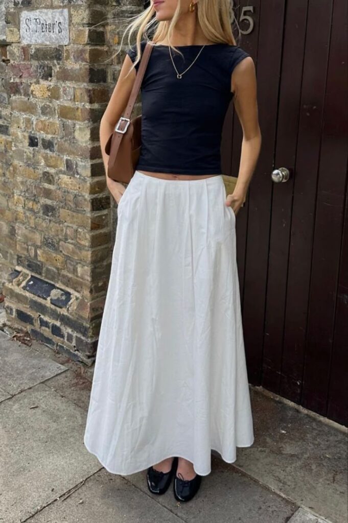 Stylish woman wearing a black top and white maxi skirt, standing against a brick wall in a chic urban setting.