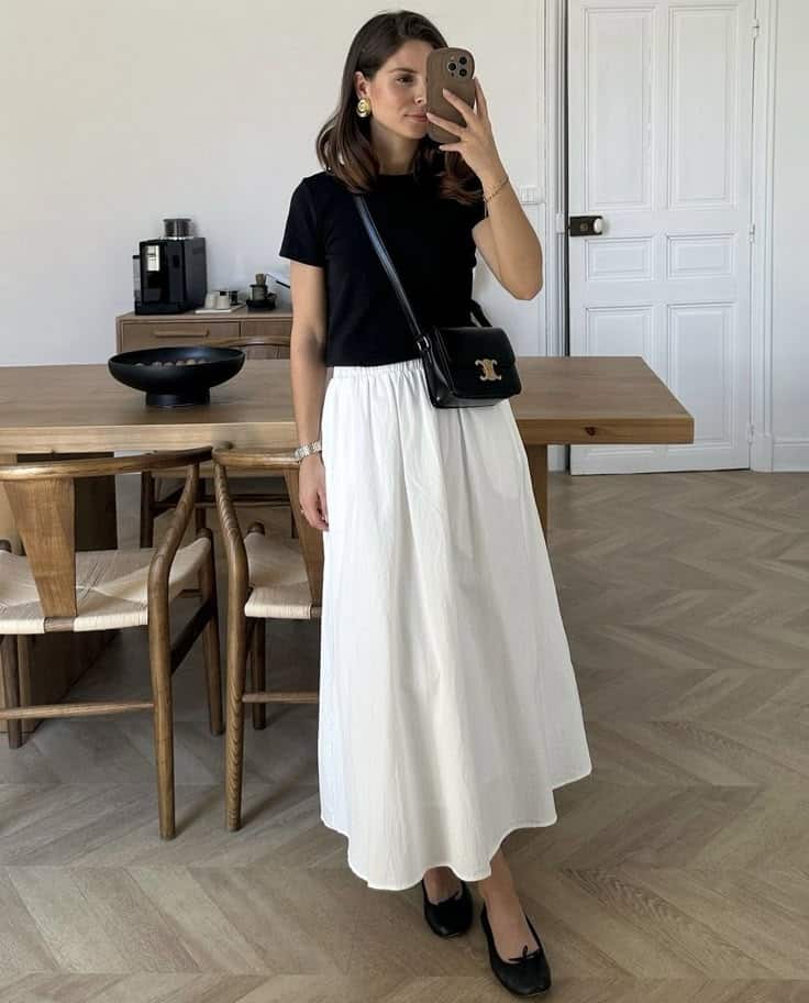 Woman in chic black top and white skirt taking a mirror selfie, stylish home decor in background.