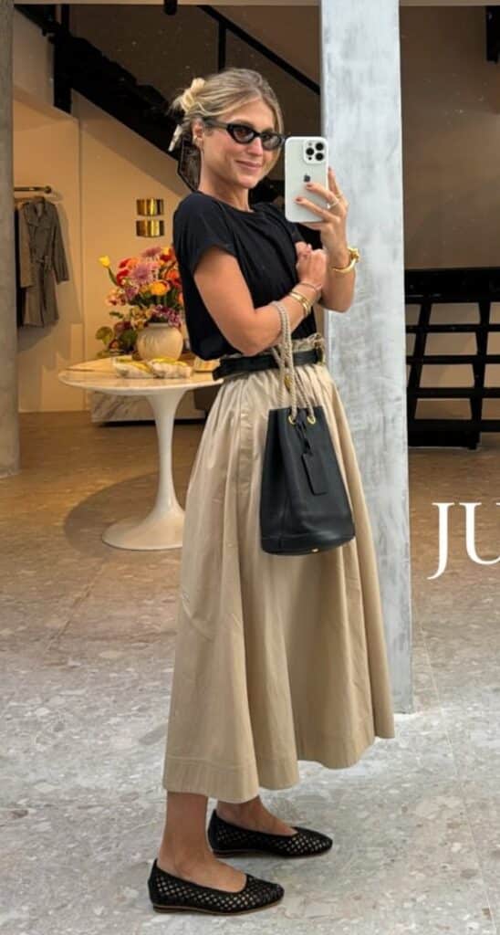 Stylish woman in black top and beige skirt posing for a mirror selfie with a chic black handbag.