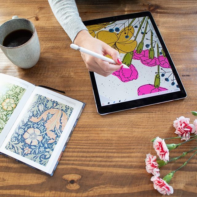 Person digitally illustrating flowers on a tablet next to a design book, coffee, and pink carnations on a wooden table.