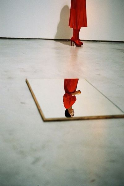 Woman in red dress and heels reflected in a floor mirror on white concrete.