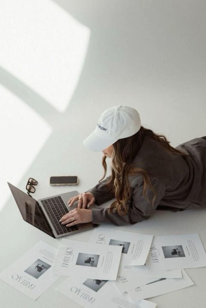Woman in white cap using laptop, surrounded by papers and phone on white surface. Relaxed and focused workspace scene.