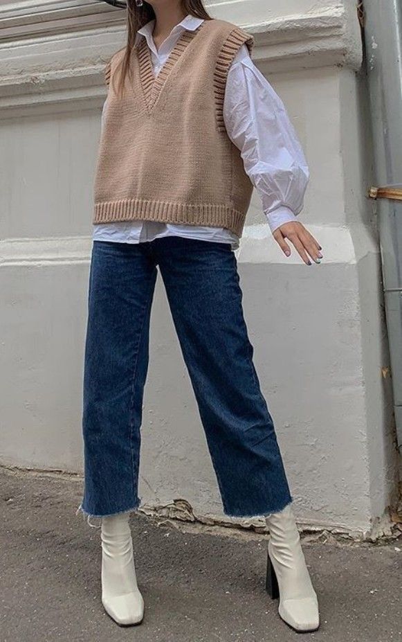 Fashionable outfit with beige knit vest, white shirt, wide-leg jeans, and white boots against a neutral background.