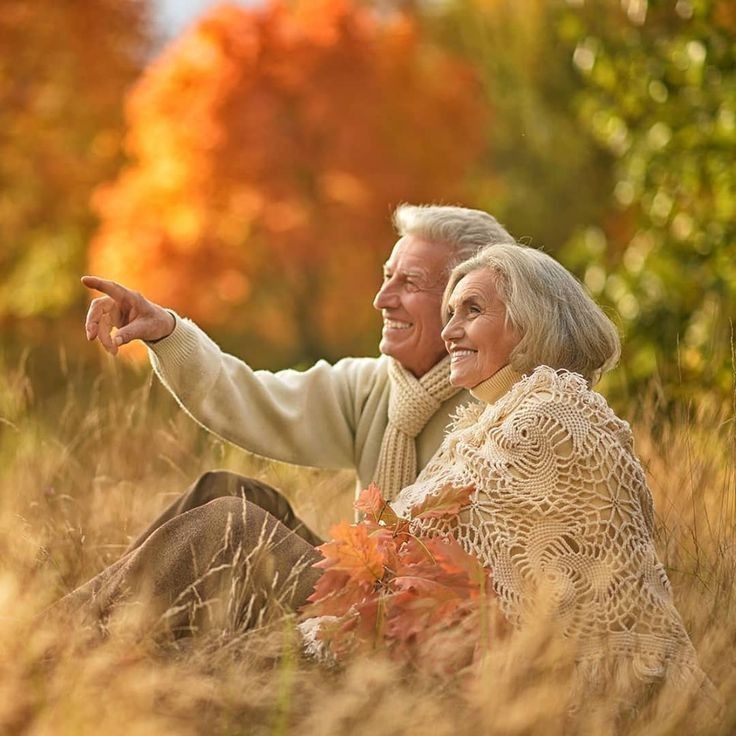 Elderly couple enjoying autumn, sitting in a field with colorful foliage, dressed warmly, sharing a joyful moment.