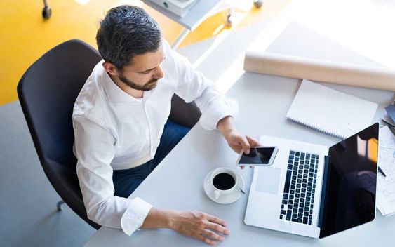 Man working at a desk with a laptop and coffee, using a smartphone, top view.