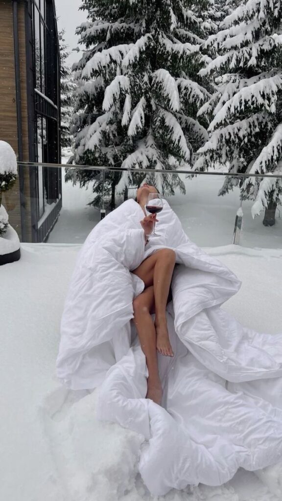 Cozy relaxation in snowy landscape, woman wrapped in blanket with wine glass on snowy balcony amidst trees.