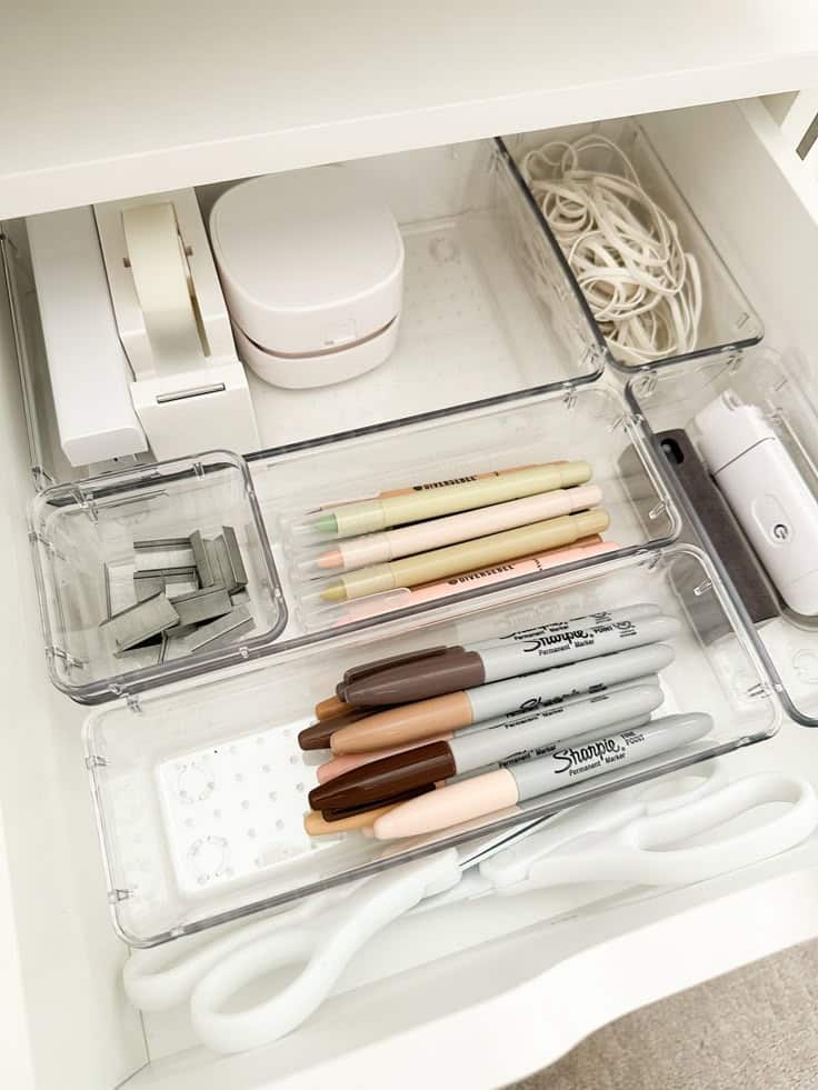 Organized desk drawer with pens, markers, tape, rubber bands, and scissors in clear storage containers.