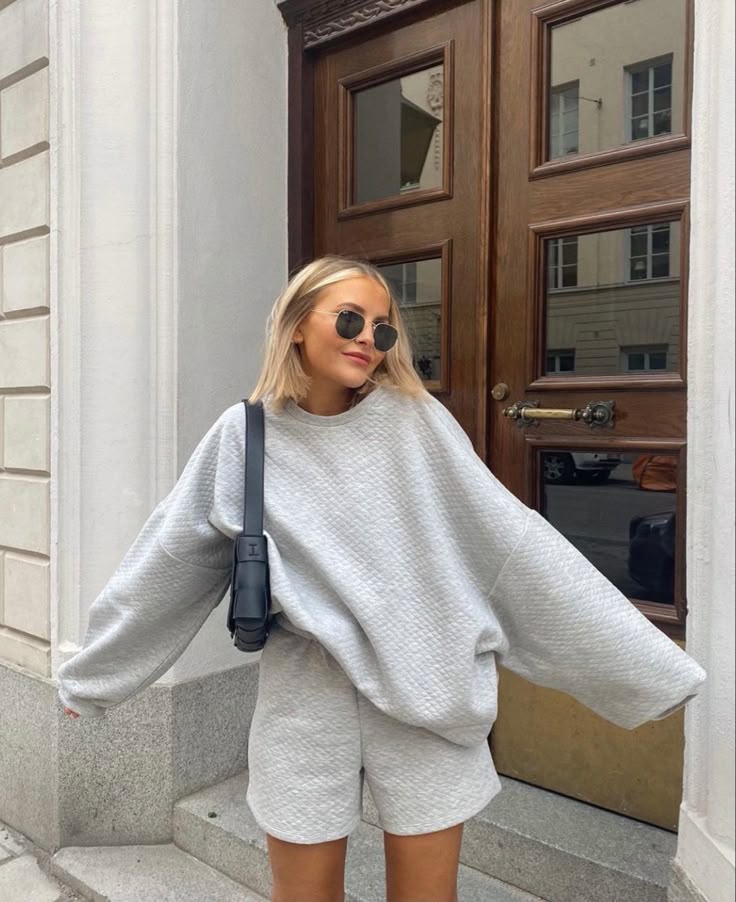 Woman in a gray matching outfit with sunglasses stands outdoors, leaning casually against a wooden door.