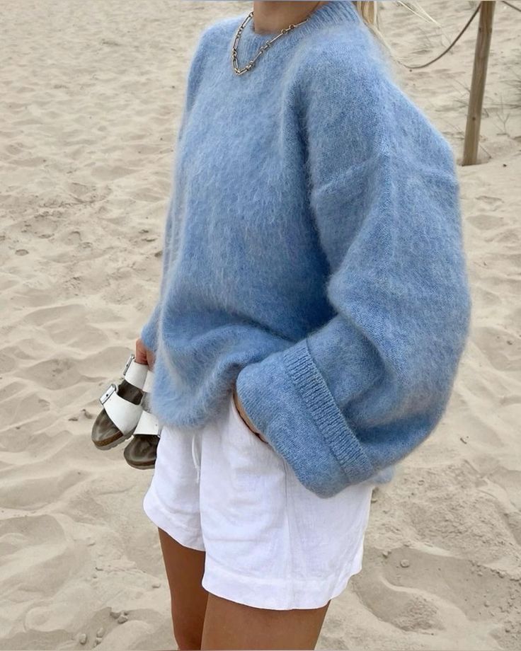 Person in blue sweater and white shorts holding sandals on a sandy beach.