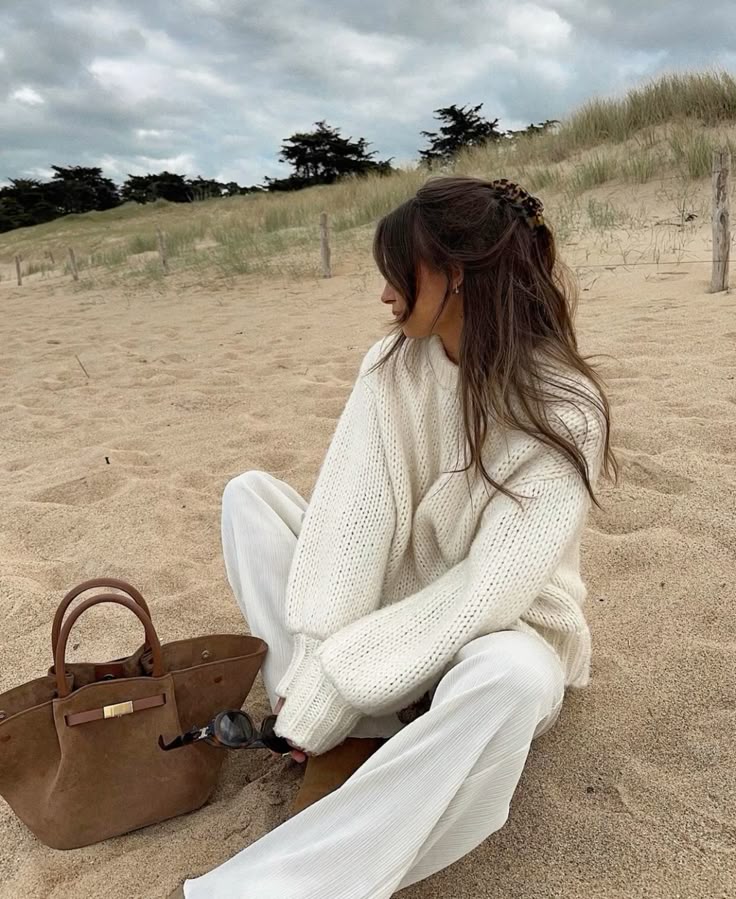 Woman in cozy sweater sits on sandy beach with stylish bag and sunglasses, overcast sky and dune grass in the background.