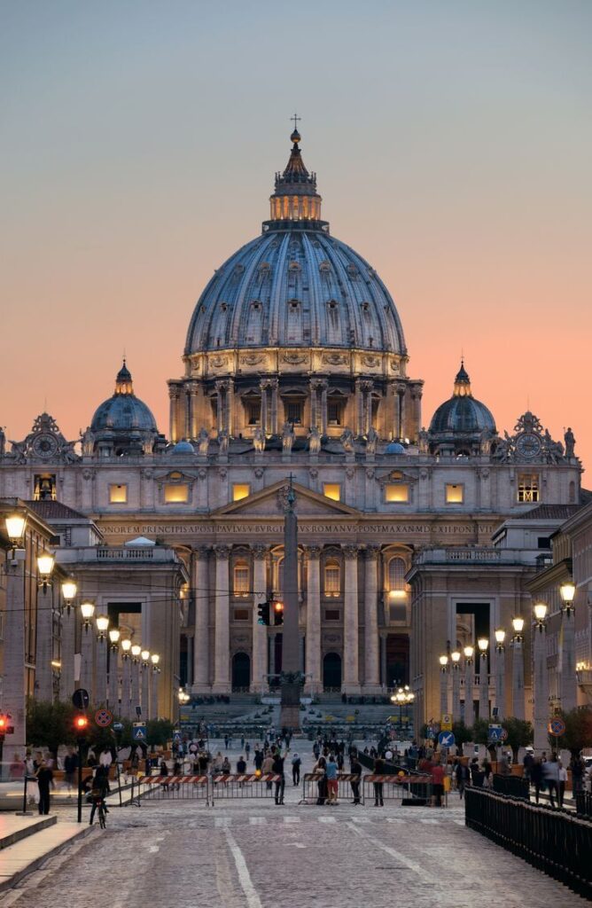 St. Peter's Basilica at sunset, Vatican City, with illuminated dome and lively foreground activity.