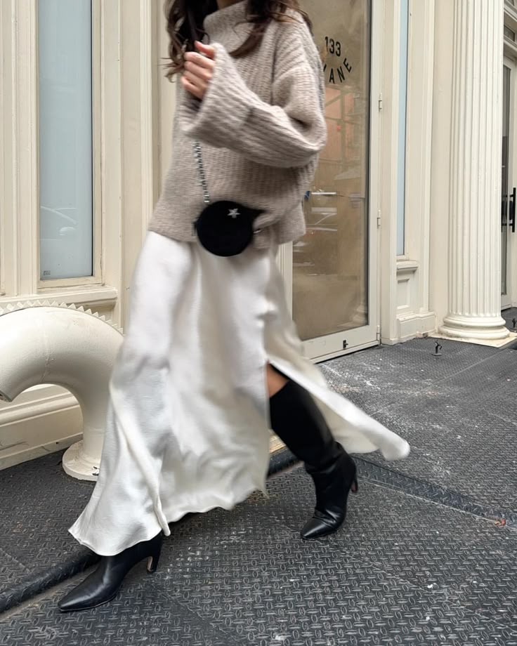 Woman in white skirt, beige sweater, and black boots walking by a building. Stylish street fashion moment.