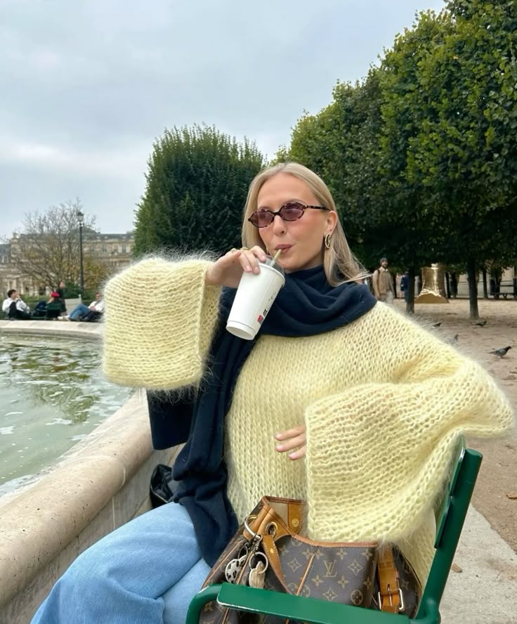 Woman wearing sunglasses and a fluffy yellow sweater drinks from a cup while sitting by a park fountain.