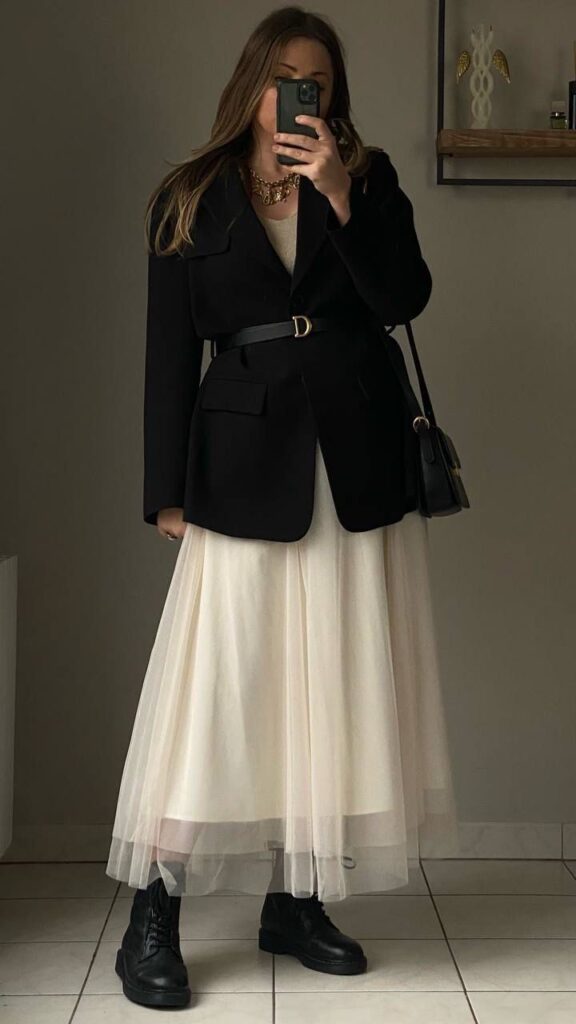 Woman in stylish blazer and tulle skirt, wearing combat boots, takes mirror selfie in chic outfit.
