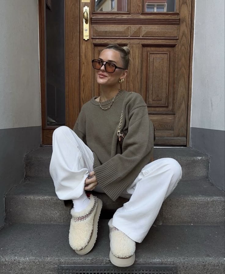 Fashionable woman in oversized sweater and white pants sitting on steps, wearing sunglasses and slippers in front of door.